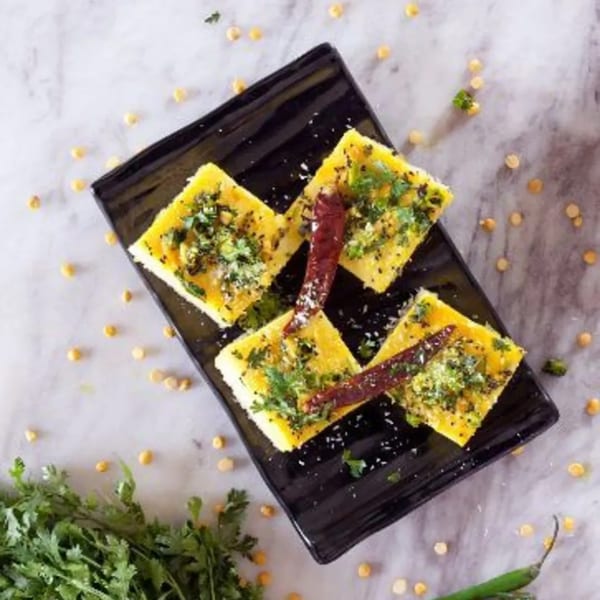 Four pieces of Kesar's Dhokla, garnished with herbs and red chilies, on a black plate.