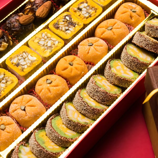 A red box filled with rows of colorful Indian sweets, including yellow squares, orange rounds, and green rolls.