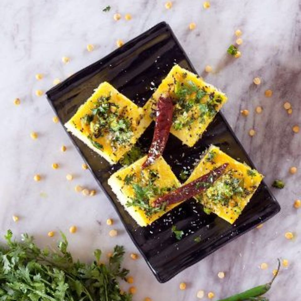 Four pieces of Kesar's Indian Dhokla, garnished with herbs and red chilies, on a black plate.