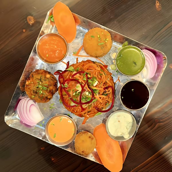 A Kesar's Indian Thali with noodles, fried patties, sauces, and sliced onions on a metal tray.