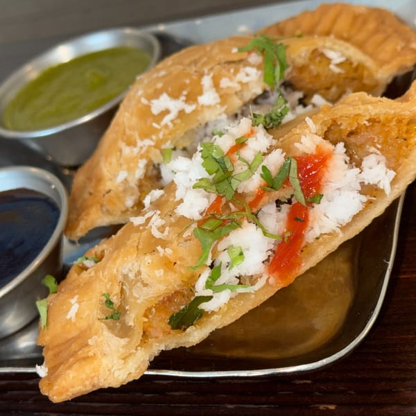 Indian samosas topped with white shredded coconut, red sauce, and green herbs, served with two dipping sauces.